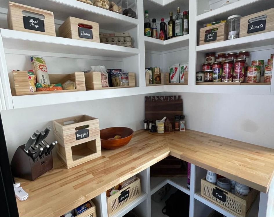 A neatly organized pantry with labeled wooden boxes, canned goods, spices, oils, jars, and a knife block on wooden countertops. Shelves hold baskets labeled snacks, drinks, and staples.
