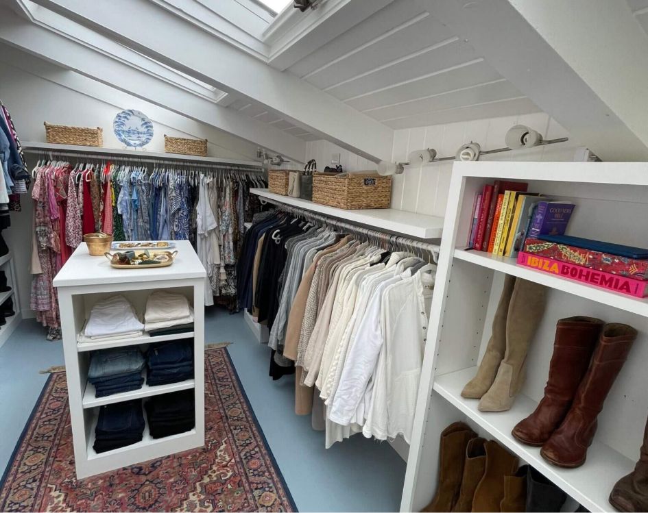 A bright walk-in closet with hanging clothes, shelves holding boots, books, woven baskets, and folded pants. An island with folded jeans sits atop a patterned rug under skylights.