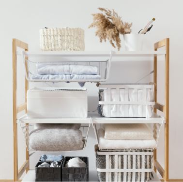 White shelving unit with baskets, folded towels, and storage boxes. Decorative vase with dried grass and a woven basket sit on the top shelf. Shelves and baskets are neatly organized.