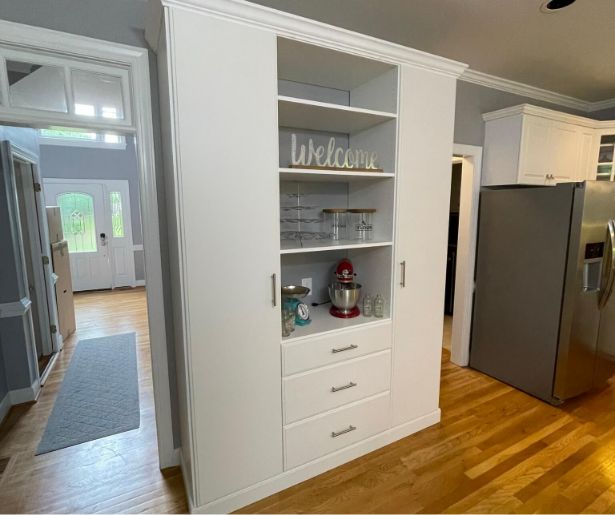 A white built-in kitchen cabinet with shelves, drawers, glassware, and a red stand mixer. A decorative Welcome sign sits on the top shelf. Hardwood floors and modern appliances are visible.