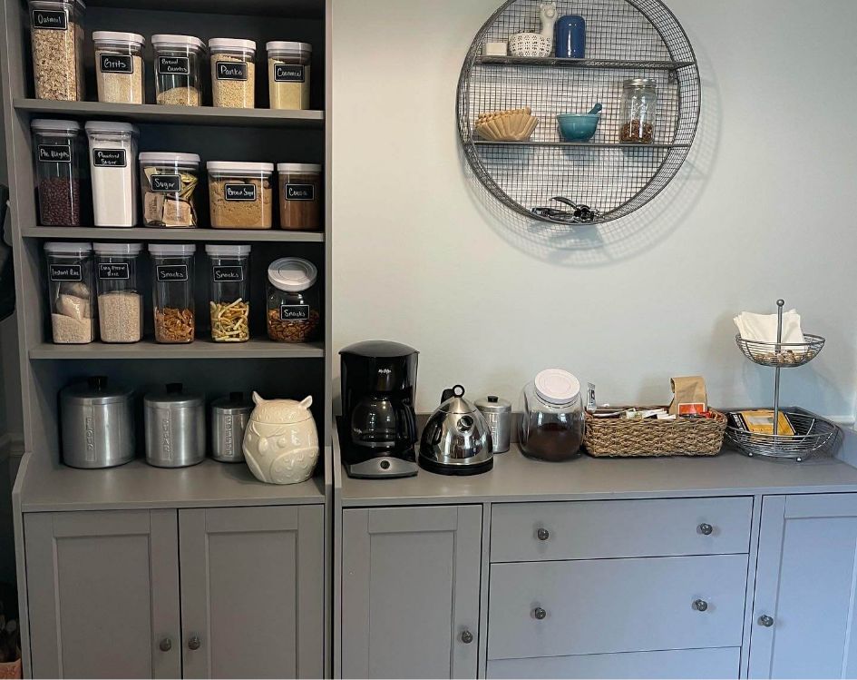 A kitchen counter with labeled containers of dry goods, a coffee maker, kettle, canisters, and a wire shelf with cups and bowls on the wall above. Everything is neatly organized on gray cabinets.