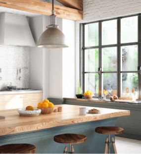 Bright kitchen with a wooden island, three stools, pendant lighting, large windows, and bowls of oranges on the counter and windowsill. Light streams in, highlighting fresh, modern decor—perfect for hosting gatherings or exploring new recipes inspired by our services showcased in the Mega Menu.