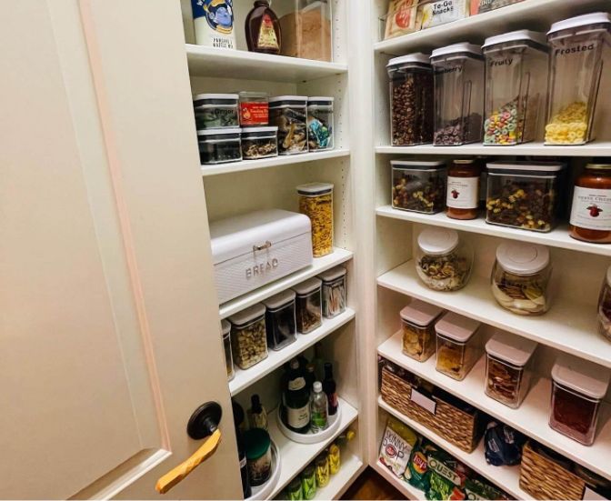 An organized pantry with shelves holding labeled containers of dry goods, jars, baskets, canned foods, and a white bread box. The shelves are neatly arranged and fully stocked.