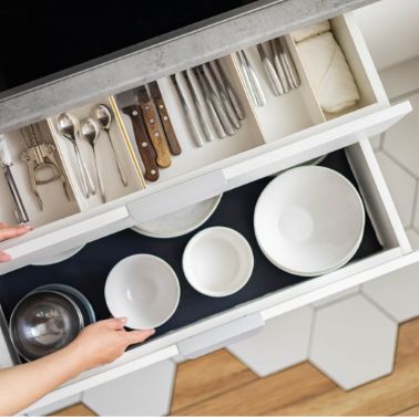 A person opens two organized kitchen drawers: the top contains cutlery and knives, while the bottom has neatly arranged white bowls and plates. The floor features large hexagonal tiles.