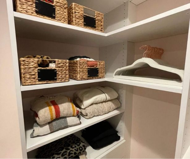 Organized closet shelves with woven baskets, folded sweaters, and a stack of white hangers. Sweaters include neutral colors and a striped pattern. Shelves and walls are white.