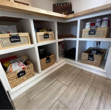 A neatly organized pantry with six labeled wicker baskets on white shelves, containing items such as snacks, rice, cups, and wellness products. The floor has beige tiles, and wooden countertops are above the shelves.