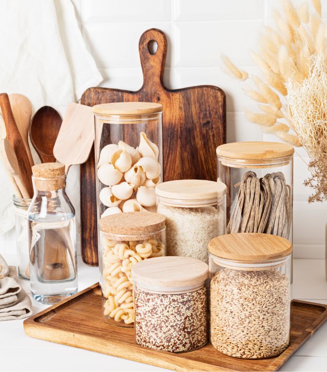 Various glass jars filled with pasta, grains, and quinoa sit on a wooden tray in a kitchen, with wooden utensils, a bottle, and a cutting board in the background.