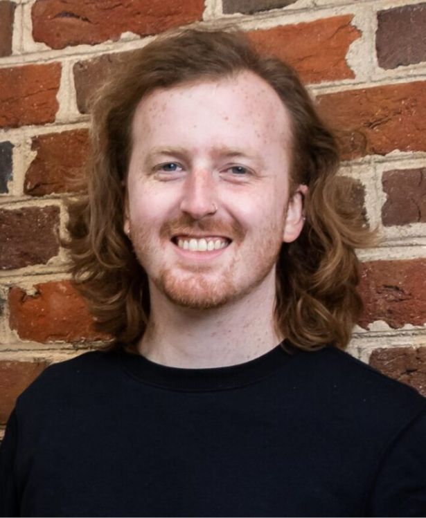 A person with shoulder-length wavy red hair and a beard smiles in front of a brick wall, wearing a black shirt.
