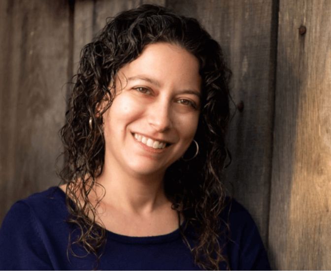 A woman with curly dark hair, wearing a navy top and hoop earrings, smiles while leaning against a wooden wall.