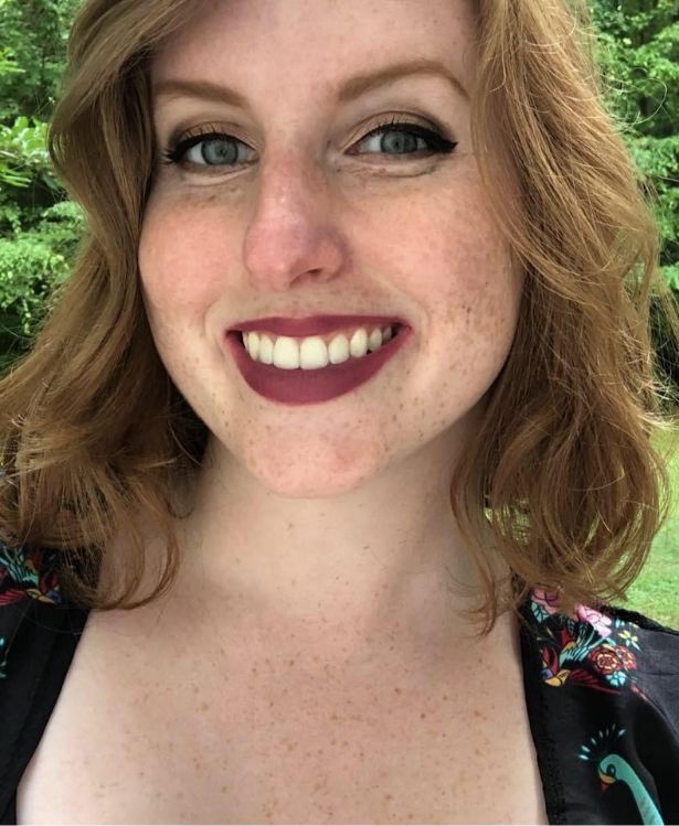 A woman with wavy, light brown hair smiles outdoors. She has fair skin, green eyes, and is wearing plum lipstick. A patterned top is visible, and lush greenery is in the blurred background.