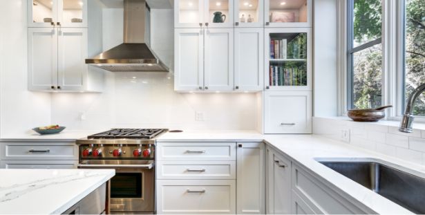 Modern white kitchen with stainless steel stove and range hood, glass-front cabinets, bookshelves, large window, and marble countertops. The sink is under the window, and recessed lighting brightens the space—perfect for organizing resources or planning meals with a mega menu of culinary ideas.