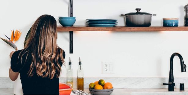 A person with long hair arranges flowers in a vase on a kitchen counter. Shelves above hold plates, bowls, and a pot. The counter also has olive oil bottles, fruit, and a black faucet—creating a scene reminiscent of the warm, inviting spaces featured in our About Us section.