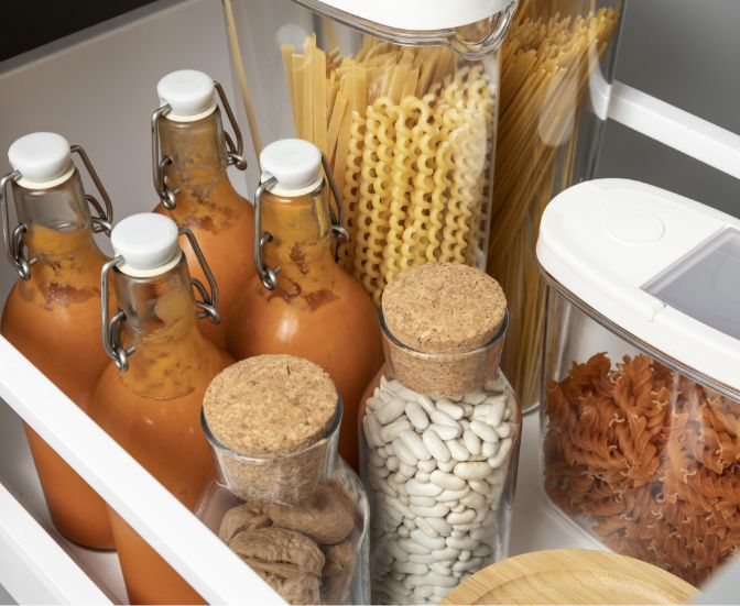 Bottles of orange sauce, glass jars with white beans and dried mushrooms, and containers of different pasta shapes are neatly organized in a white drawer.