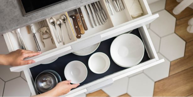A person opens two kitchen drawers, one containing neatly arranged bowls and plates, and the other filled with organized cutlery, knives, and kitchen utensils—each drawer acting as a practical mega menu of kitchen resources. The drawers are white and the floor has hexagonal tiles.