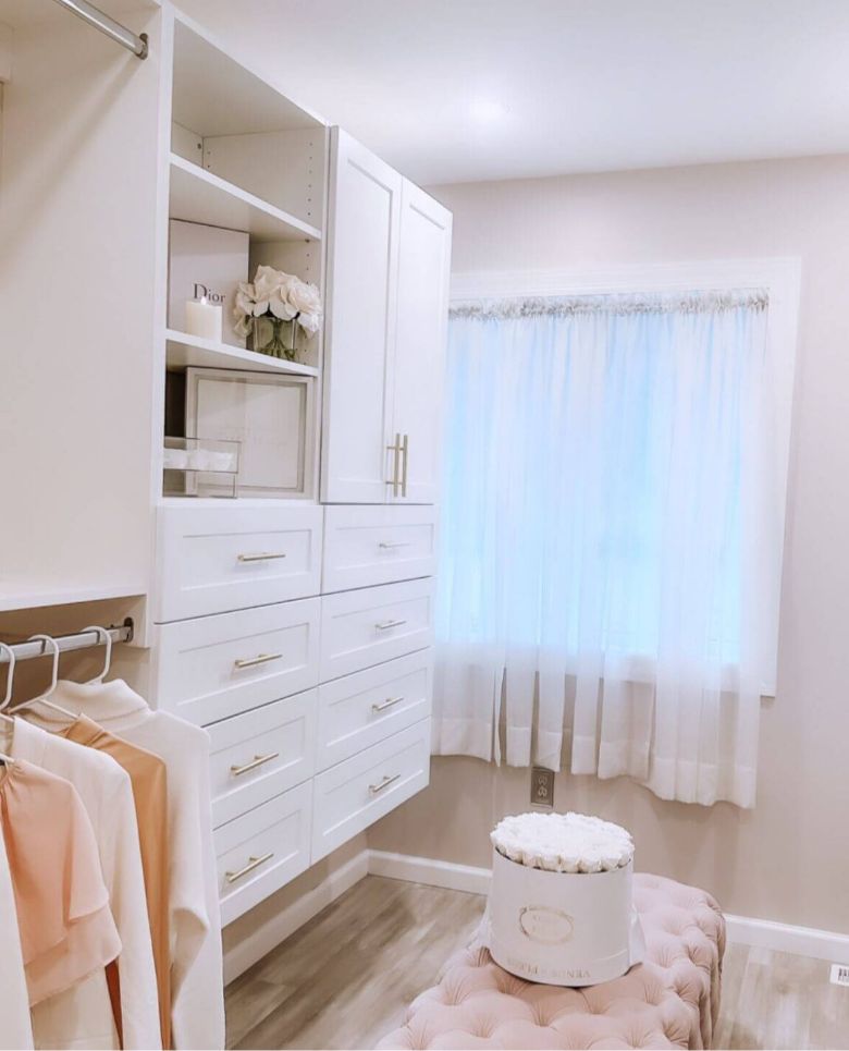 A bright, elegant closet with white cabinets, gold handles, clothes hanging on a rack, a pink tufted ottoman, and a round flower box beside a sunlit window with sheer white curtains.