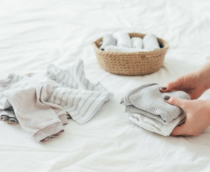Hands folding soft, neutral-colored clothes on a white bed. A basket with rolled clothes sits in the background.