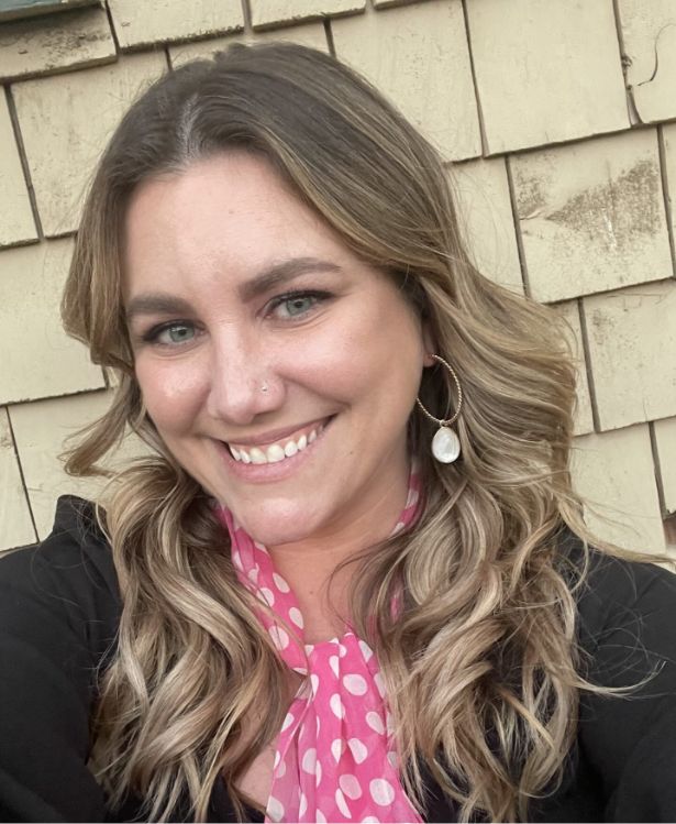 A woman with wavy blonde hair and a pink polka dot scarf smiles at the camera, standing in front of tan shingles.