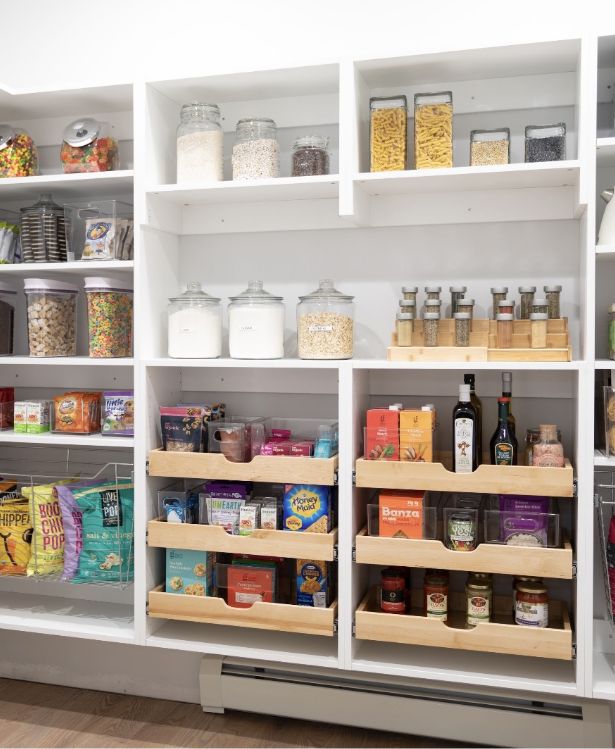 A neatly organized pantry with shelves holding jars of dry goods, baskets, spices, pasta, cereal, oils, and various snacks, all arranged in a bright, white shelving unit.