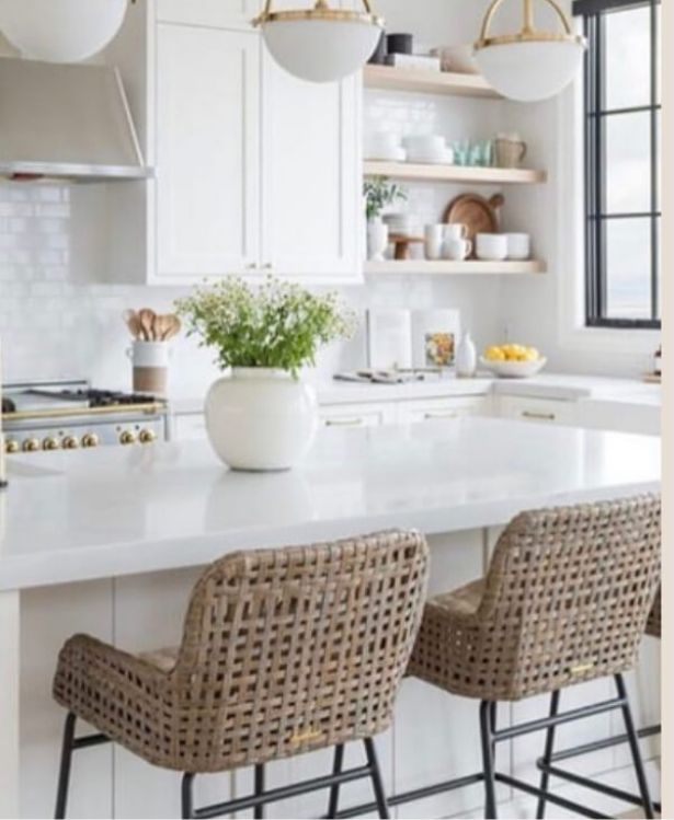 Modern kitchen with white cabinets, a white island, woven barstools, open wood shelves holding dishes, a vase of flowers on the counter, and large windows letting in natural light.