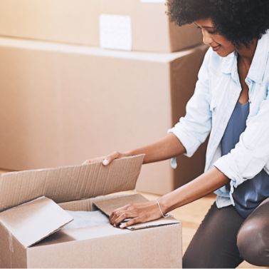 A woman kneels on the floor and packs items into a cardboard box, surrounded by other boxes, suggesting she is moving or organizing.