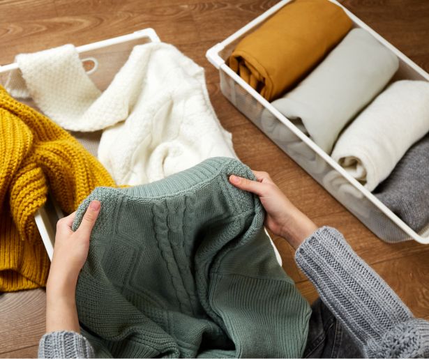 A person folds a green sweater, with neatly rolled sweaters in white bins nearby on a wooden floor. The scene shows organized knitwear in various colors, including yellow, white, brown, and gray.
