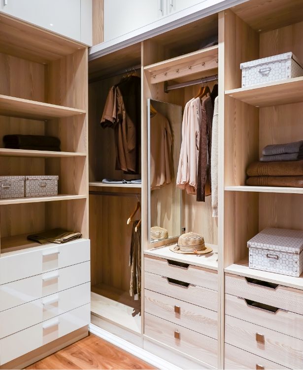 A modern wooden walk-in closet with hanging clothes, shelves, drawers, storage boxes, and a straw hat. The closet is neatly organized with neutral and brown tones.
