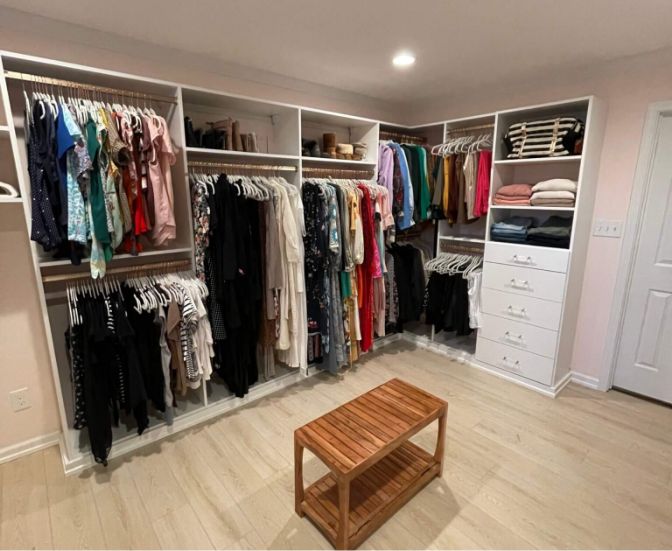 A neatly organized walk-in closet with hanging clothes, folded items on shelves, several drawers, and a wooden bench in the center on a light wood floor.